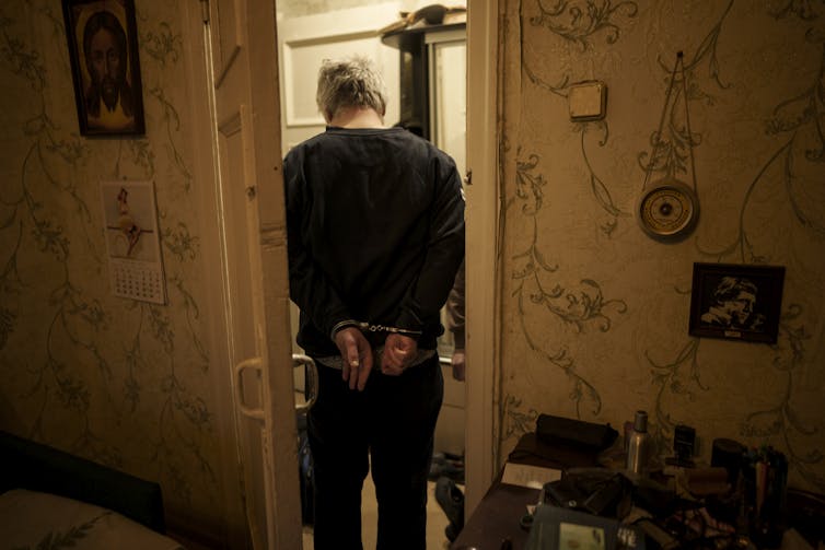 A man in handcuffs in a darkened apartment with his back to the camera.