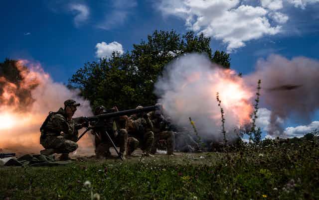 Soldiers crouch, their hands over their ears, as they shoot a large gun. Orange and pink puffs of smoke surround them.