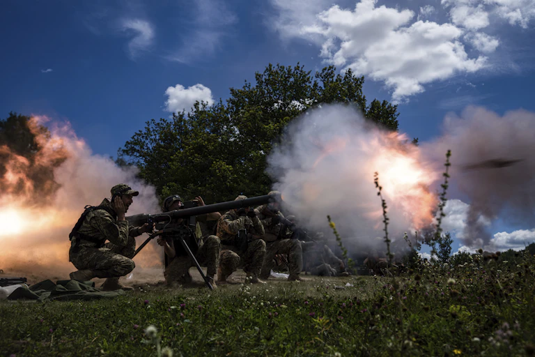Soldiers crouch, their hands over their ears, as they shoot a large gun. Orange and pink puffs of smoke surround them.