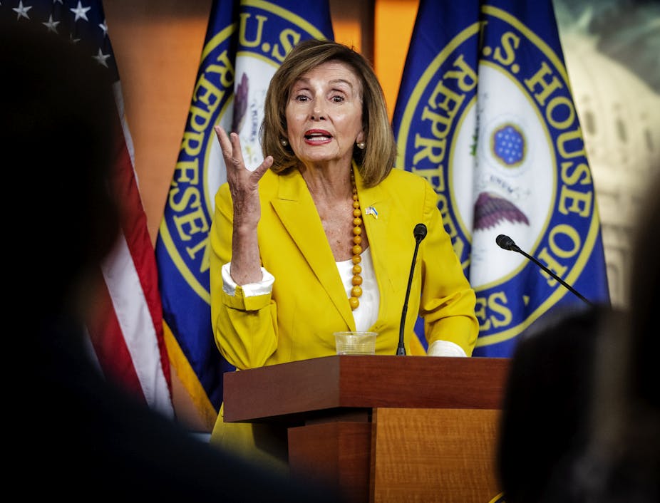 House Speaker Nancy Pelosi in a yellow blazer gesticulates to the camera with flags of the House of Representatives behind her.