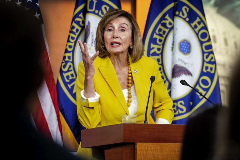 House Speaker Nancy Pelosi in a yellow blazer gesticulates to the camera with flags of the House of Representatives behind her.