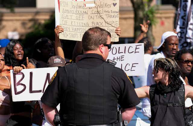 A police officer stands with his back to the camera as he watches a crowd of protesters.