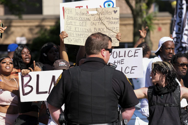 A police officer stands with his back to the camera as he watches a crowd of protesters.
