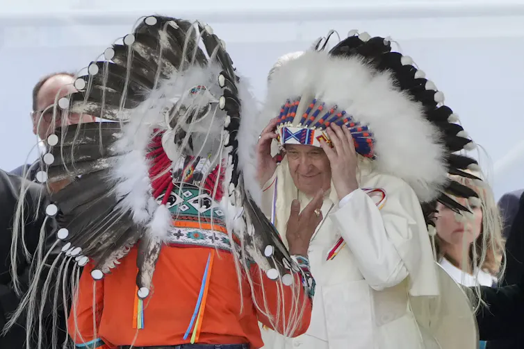 A man in a white robe straightens and Indigenous headdress on his head.