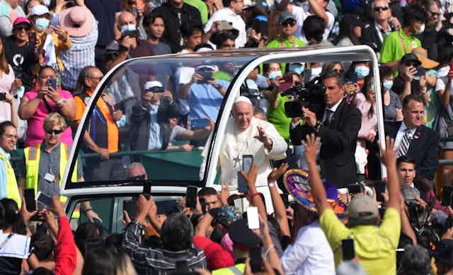An elderly man in white robes is seen waving at throngs of people from a vehicle that surrounds him in protective glass.