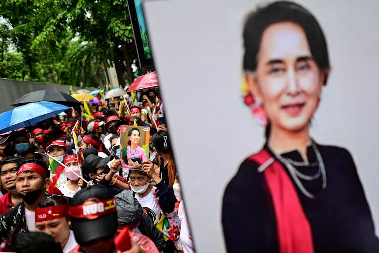 Protesters in Bangkok hold photos of Aung San Suu Kyi as they march through the streets, with umbrellas and flags.