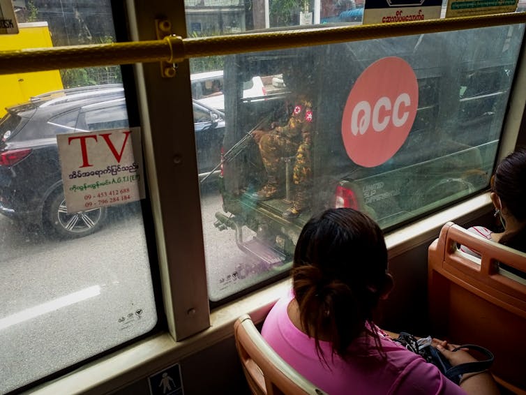 An assault weapon and the uniformed legs of a soldier are visible in the back of a truck as seen through the window of a bus on a busy city street.