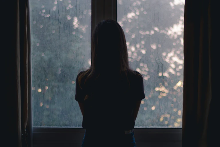 A woman shrouded in darkeness look out of an open window.
