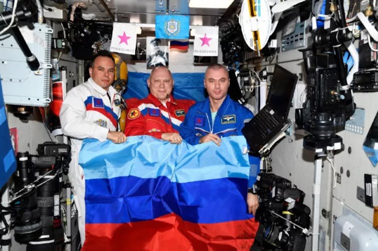 Three people holding a teal, blue and red flag aboard the ISS.