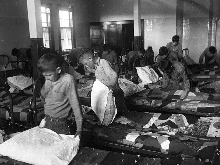 A black and white photo shows rows of boys making simple cot beds.