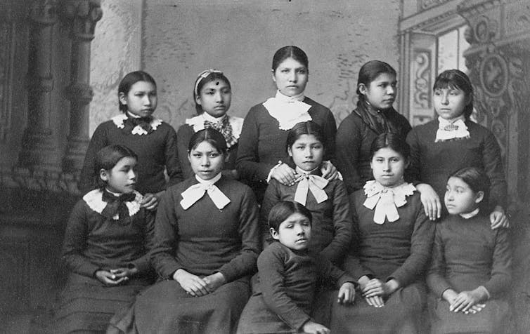 A black and white photo shows two rows of girls in black dresses with white collars.