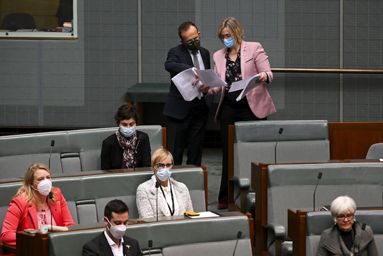 Man and women in masks peer at document, others seated nearby