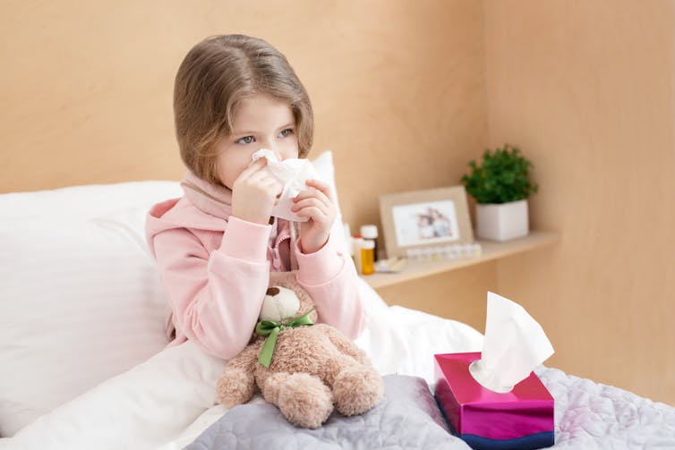 A young girl is tucked in bed with a cold. She blows her nose with a tissue.