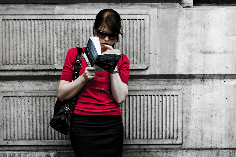 Una mujer lee un libro apoyada en una pared.