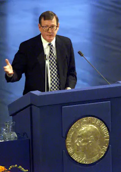David Trimble, wearing a suit and glasses, speaking at a blue podium with the Nobel crest on the front