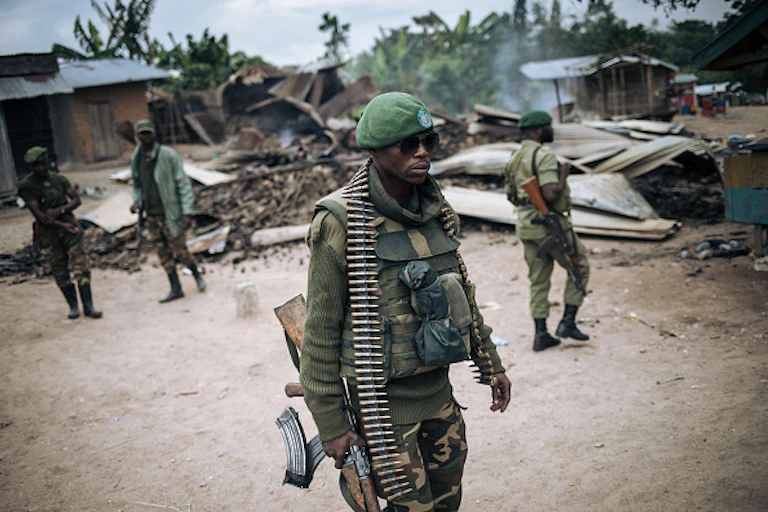 A man in camouflage military uniform, carrying a weapon, and others like him in the background, outdoors in a village with collapsed buildings