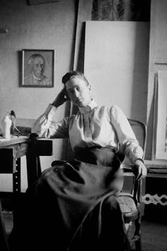 The painter Hilma af Klint sits on a chair and looks at the camera, wearing a white blouse and a long skirt.