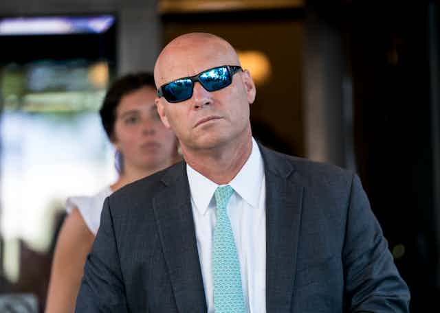 A bald man wearing sunglasses and suit and tie walks, expressionless.
