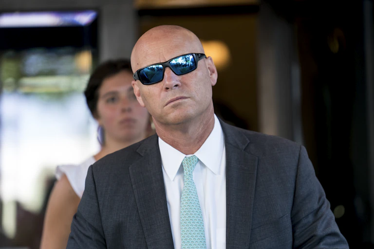 A bald man wearing sunglasses and suit and tie walks, expressionless.