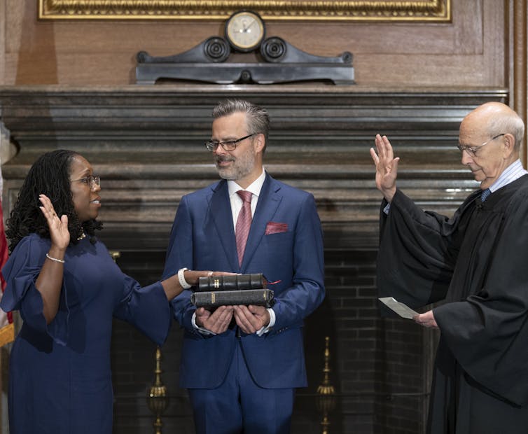 An elderly white man wearing glasses holds his hand up to face a middle aged Black woman while another man holds a Bible out for her to put her other hand on.