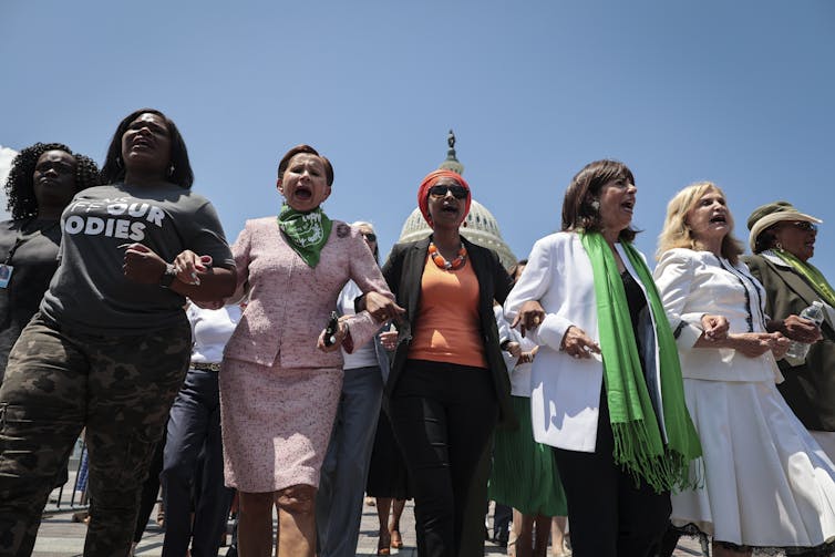 Women in a row link arms and stand in front of the U.S. Capitol