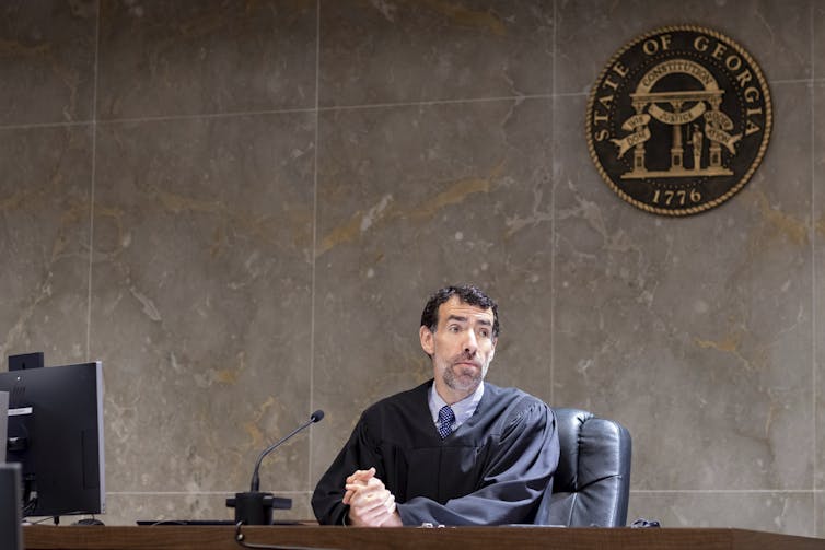 A bearded man in judge's robes sitting at a large desk, with the state seal of Georgia on the wall behind him.