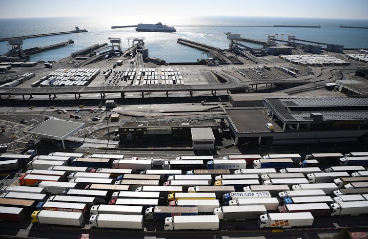 A long line of heavy trucks against the backdrop of harbor infrastructure and the sea