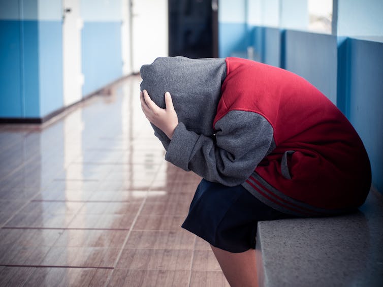 A young person wearing a hoodie is sitting alone in a hallway, holding their head in their hands.