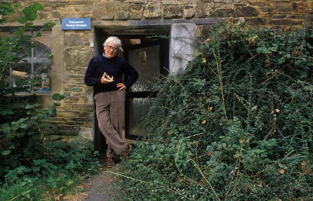 A man with white hair and glasses stands outside a door marked 'exponential dilution chamber' surrounded by vegetation.