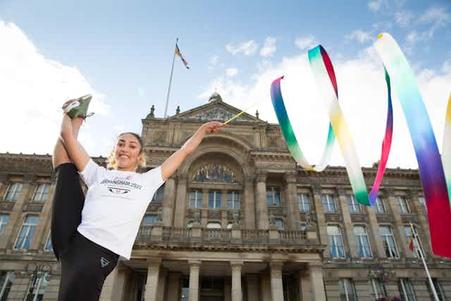 A gymnast poses with a multicoloured ribbon in front of an old building.
