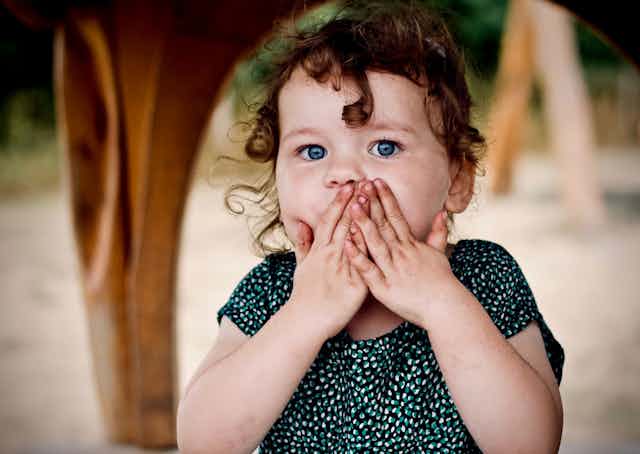 Una niña pequeña se tapa la boca con las manos con los ojos muy abiertos.