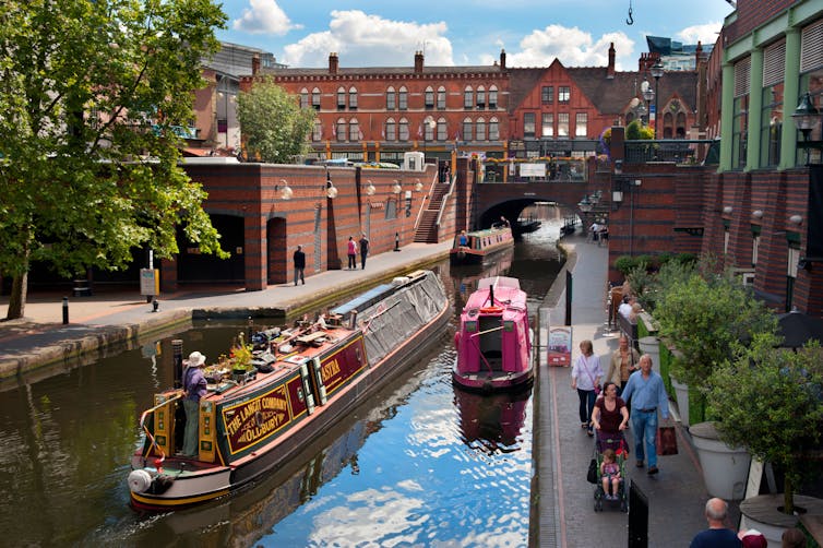 People walking alongside a canal, surrounded by dark red brick buildings while canal boats float on the canal.