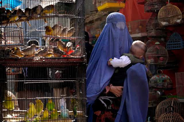 A woman in a blue burka holds her baby as she walks past cages of colourful birds.