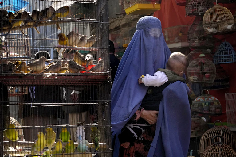 A woman in a blue burka holds her baby as she walks past cages of colourful birds.