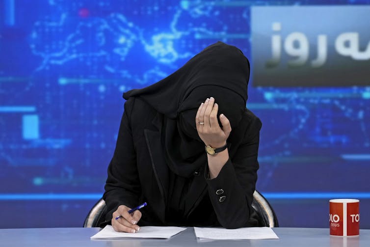 A woman in a black burka on a TV set bows her head.