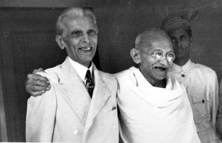 Two men -- one dressed in a white suit and another with a white shawl draped over him -- standing next to one another and laughing.
