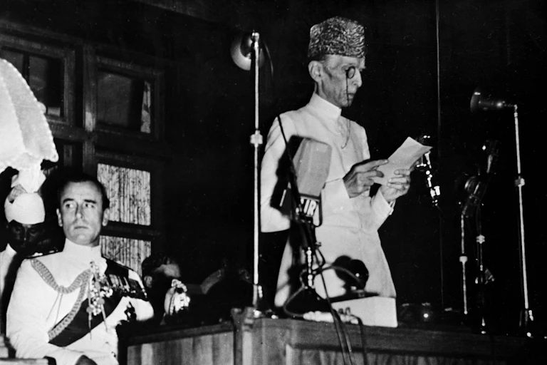 Pakistan's governor general reading a speech from the podium while the outgoing governor general of India, Lord Mountbatten looks on.