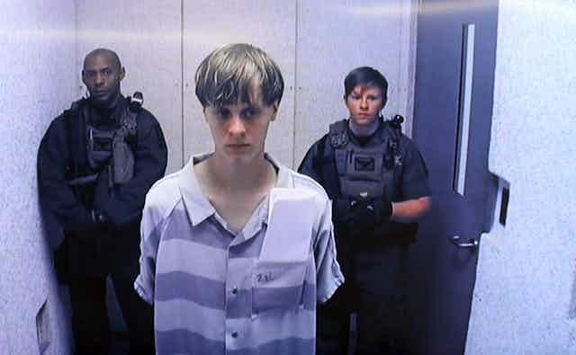 A young white man appears on a television screen wearing a purple and white prison uniform, with two guards behind him