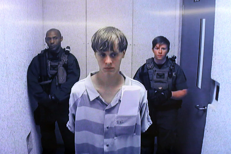 A young white man appears on a television screen wearing a purple and white prison uniform, with two guards behind him