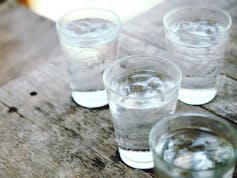 Four clear glasses of water with ice floating in them sit on a wooden surface.