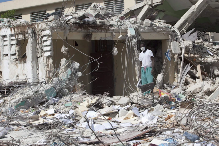 A person wearing medical scrubs and a surgical mask stands in a the rubble of a destroyed building