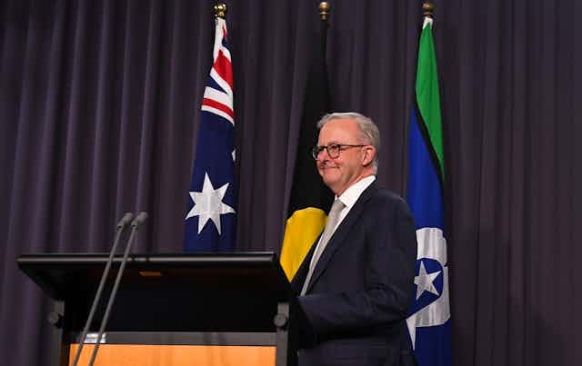 Anthony Albanese, a white man in a suit and glasses, stands at a podium in front of the Australian, Aboriginal and Torres Strait Islander flags.