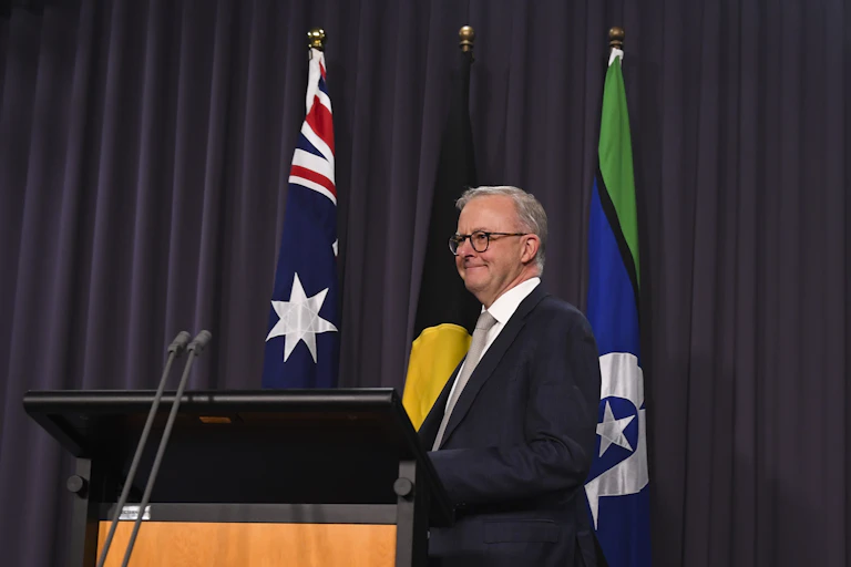 Anthony Albanese, a white man in a suit and glasses, stands at a podium in front of the Australian, Aboriginal and Torres Strait Islander flags.