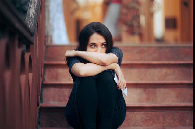 A young woman sitting on a set of stairs hugs her knees, looking anxious.