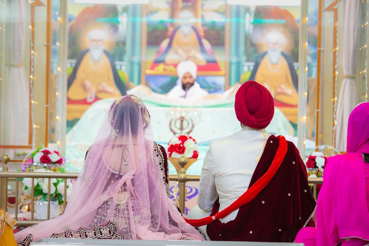 A couple in Sikh wedding clothes seen from the back during a ceremony.