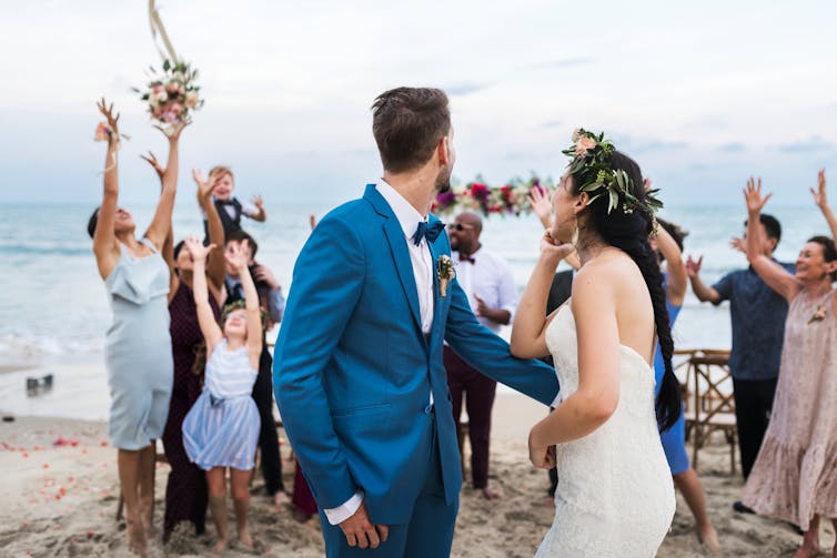 A couple look back at guests scrambling for a bouquet on a beach.