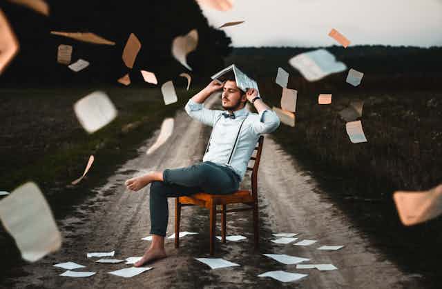 Un hombre sentado en una silla en un camino rural se tapa la cabeza con un libro mientras llueven hojas de papel.