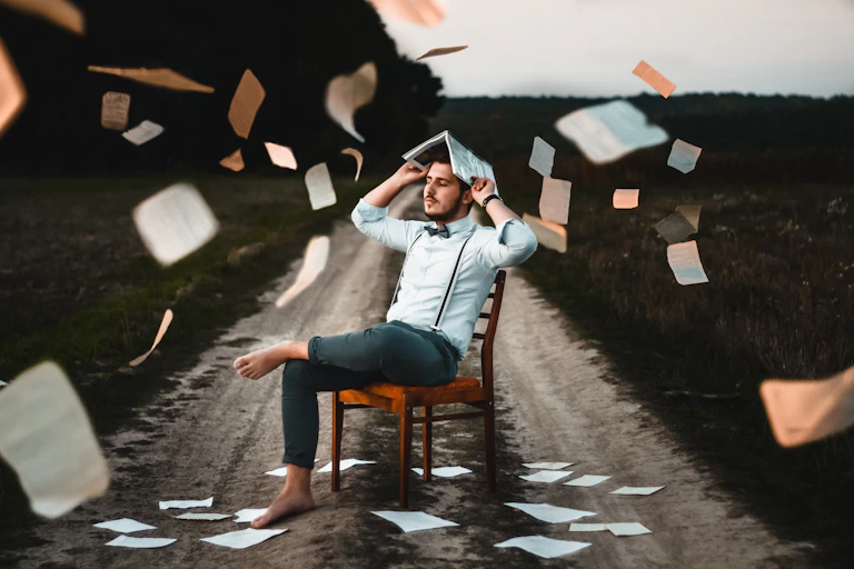 Un hombre sentado en una silla en un camino rural se tapa la cabeza con un libro mientras llueven hojas de papel.