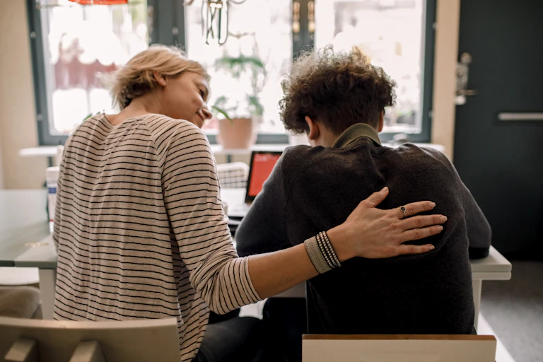 An adult puts her hand on the back of a young person sitting at a table