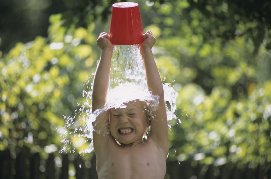 Outdoors in front of a leafy bush, a shirtless child with tightly shut eyes and a clenched smile holds a bucket upside down over their head as a gush of water splashes onto them.